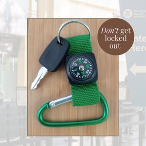 May include: A key, a black compass, and a green carabiner are attached to a green strap. The text "Don't get locked out" is in a brown circle. The background is a light brown wood grain.