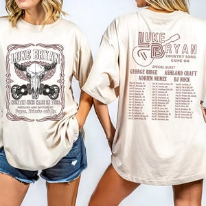 May include: Cream-colored t-shirt featuring the text "LUKE BRYAN COUNTRY SONG CAME ON TOUR" with a bull skull and guitar graphic. The back of the shirt lists tour dates and special guests George Birge, Adrien Nunez, Ashland Craft, and DJ Rock.