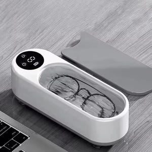 以下が含まれることがあります： タイマーとバッテリーアイコンを表示するデジタルディスプレイを備えた、白とグレーの超音波洗浄機。装置には水で満たされた開いた長方形のタンクがあり、洗浄のために黒いフレームの眼鏡が沈められています。