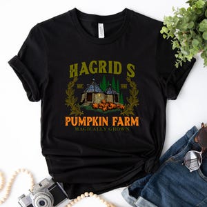 Könnte beinhalten: Schwarzes T-Shirt mit einem Grafikdruck einer kleinen Hütte mit Kürbissen und dem Text "Hagrid's Pumpkin Farm Magically Grown".