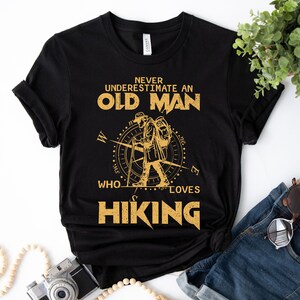 Op de afbeelding: Zwart T-shirt met een gouden grafisch ontwerp met de tekst "Onderschat nooit een oude man die van wandelen houdt". De grafiek bevat een kompas en een silhouet van een wandelaar.