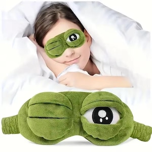 May include: A plush green sleep mask featuring a cartoon frog design with one eye closed in a wink and the other eye wide open with a black and white pupil. The mask is made of soft fabric and includes an elastic strap for a secure fit over the eyes.