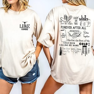 May include: Two beige t-shirts with black text and graphics. The front of one shirt says "LUKE COMBS" with a bull's head logo. The back of the other shirt has lyrics from Luke Combs songs, including "Lovin' on You", "Forever After All", "One Number Away", "Beautiful Crazy", "She Got the Best of Me", "Cold Beer Calling My Name", "Cold as You", "Beer Never Broke My Heart", and "When It Rains It Pours".