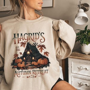 Könnte beinhalten: Ein beiger Pullover mit einem grafischen Druck eines Hauses mit Kürbissen und dem Text "Hagrid's Est. 1981 Autumn Retreat".