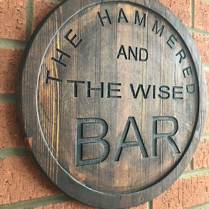May include: A circular wooden sign, stained dark brown, with the words "THE HAMMERED AND THE WISE BAR" carved into it. The sign is mounted on a red brick wall. The sign has a rustic, vintage aesthetic.
