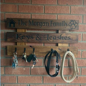 May include: A wooden key and leash holder with the text "The Morgan Family" and "Keys & Leashes". The holder has hooks for keys and leashes, and is mounted on a brick wall. A paw print and house graphic are also visible.