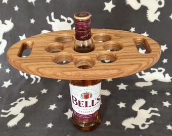 May include: A wooden serving tray with six circular cutouts and two handles. A bottle of Bell's Original Scotch Whisky supports the tray, with a small bottle of liquor in one of the holes. The tray is made of light brown wood.