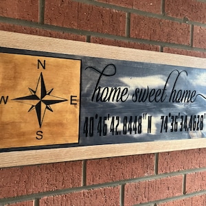 Op de afbeelding: Houten bord met een kompasroos en de tekst "Home sweet home". Coördinaten worden onder de tekst weergegeven. Het bord is omlijst met licht hout en bevestigd aan een bakstenen muur.