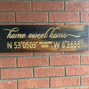 以下が含まれることがあります： 「Home sweet home」の文字が筆記体で書かれた長方形の木製サイン。その下に座標と2009年が記されています。レンガの壁に取り付けられています。