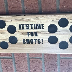 以下が含まれることがあります： 「IT'S TIME FOR SHOTS!」の黒い文字が入った木製のショットグラスホルダー。ホルダーには切り抜かれたハンドルと、ショットグラス用の円形の黒いインサートが付いています。木材は自然な明るい色の木目です。