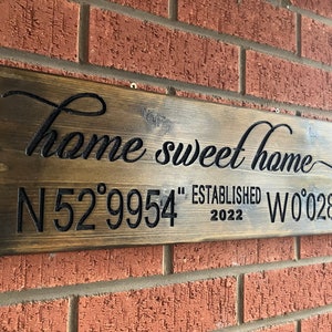 以下が含まれることがあります： 「home sweet home」の文字が筆記体で書かれた木製の看板。その下に座標と2022年が表示されています。レンガの壁に取り付けられています。