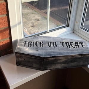 May include: A grey wooden coffin-shaped box with the words "TRICK OR TREAT" carved into the lid. The box is placed on a white surface near a window. The design suggests a Halloween theme.