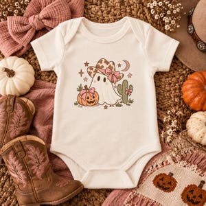 May include: A cream-coloured short-sleeve baby bodysuit featuring a graphic of a ghost wearing a brown cow-print cowboy hat with a pink bow, standing next to a jack-o'-lantern and a cactus, both adorned with small pink bows.