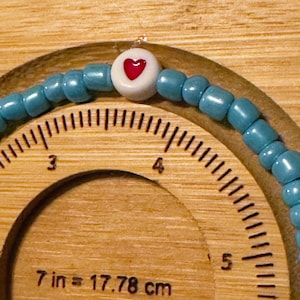 May include: A beaded bracelet with turquoise beads and a white bead featuring a red heart. The bracelet is displayed on a wooden surface with the text "7 in = 17.78 cm".