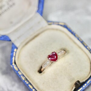 Peut inclure: Bague en or avec un rubis en forme de cœur, présentée dans un écrin bleu. La bague présente une pierre précieuse rouge vif et de petites pierres d'accent sur la bande. L'écrin a un intérieur blanc et un extérieur bleu décoratif.