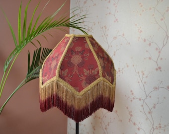 Puede incluir: Una pantalla de lámpara roja y dorada con un patrón floral y un ribete de flecos. La pantalla de la lámpara está hecha de tela y tiene un borde festoneado.