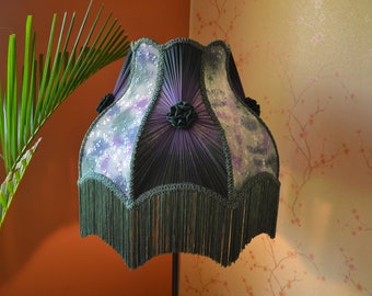 May include: A bell-shaped lampshade featuring alternating panels of dark purple pleated fabric and light purple floral patterned fabric. The shade is decorated with small fabric rosettes at the top and finished with a long, dark green fringe trim.