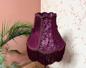 Puede incluir: Pantalla de lámpara burdeos oscuro con un estampado floral de terciopelo y ribete de flecos. La pantalla presenta un diseño plisado en un lado y una flor decorativa. El ribete y los flecos son de un tono burdeos más oscuro.