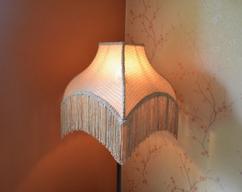 May include: A vintage-style lampshade with a woven pattern and a fringed trim. The lampshade is a warm, cream color, and the fringe is a matching light beige. The lamp is positioned in a corner, with a brown wall on one side and a floral wallpaper on the other.