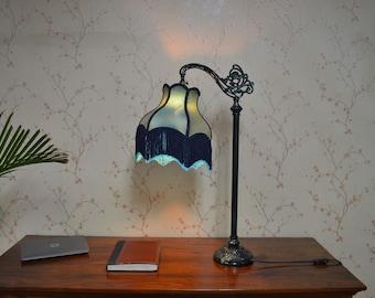 May include: A table lamp with a dark metal base featuring an ornate, curved arm and a circular stand. The lamp has a scalloped, cream-colored fabric shade trimmed with a deep navy blue fringe and a light blue decorative edge along the bottom.
