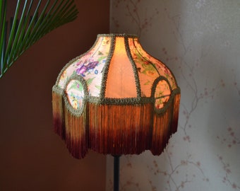 May include: A bell-shaped lampshade featuring a light pink fabric with a colourful floral and butterfly pattern. The shade is accented with gold braided trim around the circular panels and finished with a long, layered fringe in shades of gold and deep red.