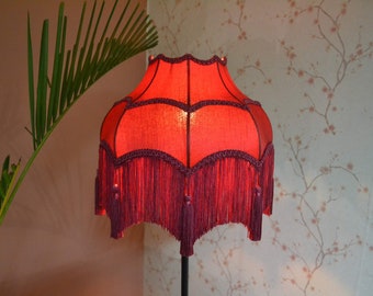 May include: A bell-shaped lampshade in a vibrant red fabric featuring a scalloped edge. The shade is adorned with dark burgundy braided trim along the seams and a long, matching fringe with decorative tassels hanging from the bottom border.