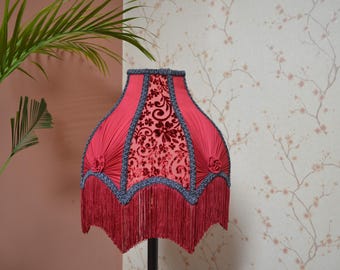 Puede incluir: Pantalla de lámpara roja con un estampado floral de terciopelo y flecos. La pantalla tiene un borde festoneado con ribete azul, sobre un fondo neutro.