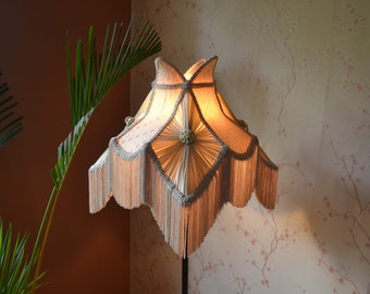 May include: A cream-colored lampshade with a fringe trim and a floral design. The lampshade is lit and casts a warm glow.