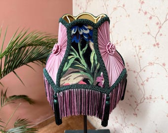 May include: A decorative lampshade with a pink pleated fabric exterior and a floral patterned interior. The shade is trimmed with green and features a fringe at the bottom. Two pink rosettes adorn the sides.