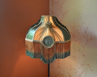May include: A bell-shaped lampshade featuring alternating panels of dark green velvet and pleated beige fabric. The shade is adorned with a central fabric rosette, decorative gold braided trim along the edges, and a long, layered fringe in orange and green.