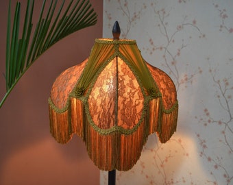 May include: A bell-shaped lampshade featuring an orange lace overlay with a floral pattern, accented by olive green fabric panels, decorative braided trim, and long, flowing orange fringe hanging from the scalloped bottom edge.
