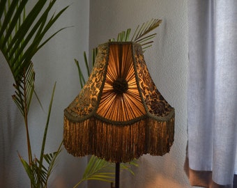 May include: A gold and green lampshade with a fringe trim. The lampshade has a floral pattern and is lit from within.