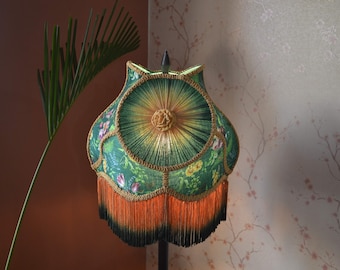 May include: A bell-shaped lampshade featuring a green fabric with a floral pattern, accented by gold braided trim and a central rosette. The bottom edge is finished with a long, dense fringe that transitions from a bright orange to a dark black.