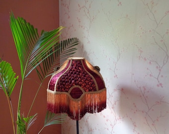 Puede incluir: Una pantalla de lámpara de color burdeos con flecos dorados y un patrón tejido. La pantalla está sobre un soporte y se encuentra frente a una pared con un patrón floral.
