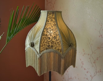 Puede incluir: Una pantalla de lámpara en forma de campana de color verde oliva apagado, con paneles alternos de tela plisada y tejido con motivos florales. La pantalla está adornada con pequeñas rosetas de tela y rematada con un largo borde de flecos.