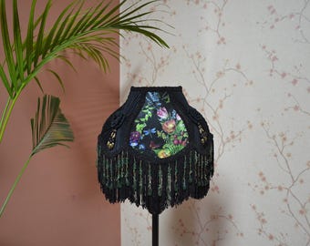 May include: A lampshade featuring a black fabric exterior with pleated sections and a central panel displaying a colorful floral and dragonfly print. The lower edge is adorned with long black fringe and dangling green crystal-like beads.