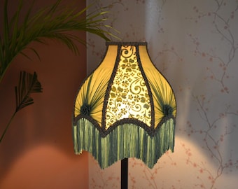 May include: A green and yellow lampshade with a floral pattern and fringe trim. The lampshade is lit and casts a warm glow.