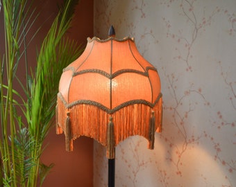 May include: A bell-shaped lampshade in a vibrant orange fabric featuring a scalloped edge trimmed with decorative gold braid and long, matching orange fringe tassels hanging from the bottom perimeter, topped with a dark pointed finial.