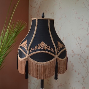 Puede incluir: Una pantalla de lámpara negra con flecos dorados y bordados dorados intrincados. La pantalla tiene un diseño clásico y elegante.