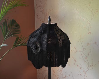 May include: A black lampshade featuring pleated fabric panels, intricate lace detailing, and decorative fabric rosettes. The lower edge is finished with a long, dense black fringe, and the top is capped with a pointed dark metal finial.