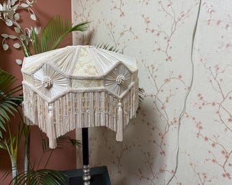 May include: Cream-colored lampshade with a decorative fringe and tassel design. The lampshade has a geometric pattern and is supported by a black and silver lamp base. The background features a floral wallpaper.