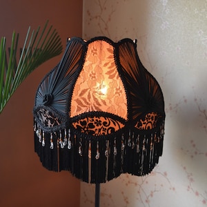 Puede incluir: Una pantalla de lámpara en forma de campana con paneles de tela negra y superposiciones de encaje intrincado, que emite un cálido resplandor naranja. La pantalla está adornada con ribetes de cordón negro, rosetas decorativas y flecos negros.