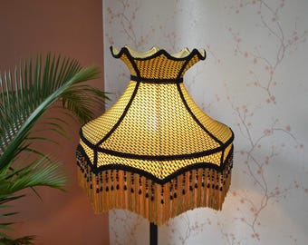 May include: A bell-shaped lampshade featuring a bright yellow woven fabric texture with a scalloped top edge. The shade is accented by thick black trim along the seams and edges, finished with a long, golden-brown fringe and black bead details.