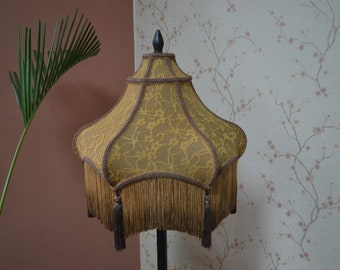 Puede incluir: Una pantalla de lámpara en forma de campana con un encaje superpuesto de color amarillo mostaza con patrón floral. La pantalla está acentuada con un borde trenzado marrón oscuro y terminada con flecos largos a juego y borlas decorativas.
