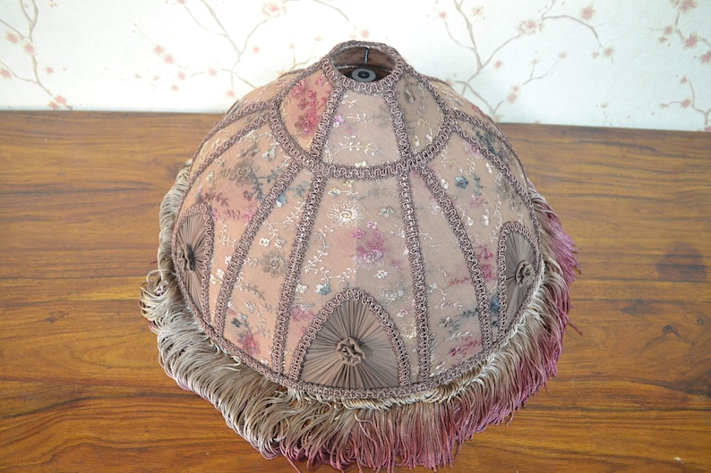 Op de afbeelding: Een ronde lampenkap met een bloemenpatroon en een bruine franje. De lampenkap is gemaakt van een delicate stof met een subtiele glans.