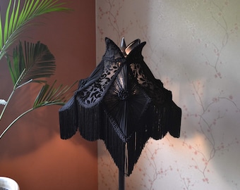 May include: A black lampshade featuring a structured, multi-paneled design with intricate lace overlays and a central diamond-shaped section. The edges are adorned with long, dense black fringe trim that hangs down from the bottom of the shade.