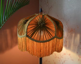 May include: A lampshade featuring a pleated, golden-orange fabric design with scalloped edges. The shade is adorned with decorative fabric rosettes at the top of each panel and finished with a long, matching golden-orange fringe trim.
