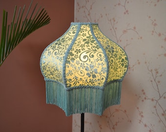 Puede incluir: Una pantalla de lámpara en forma de campana con una base de tela amarilla y un patrón repetitivo de flores y enredaderas en verde oscuro. La pantalla presenta un borde trenzado verde oscuro en las costuras y flecos largos a juego en la base.