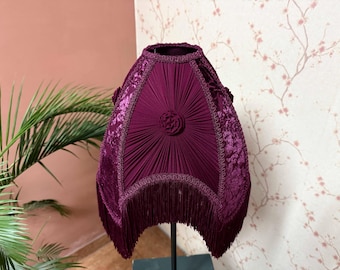 Könnte beinhalten: Ein tief burgunderfarbener Lampenschirm mit plissiertem Design und Fransen. Der Schirm hat einen verzierten floralen Besatz und eine zentrale Rosette. Der Lampenschirm ist im Vintage-Stil gehalten und ist wahrscheinlich für eine Tischlampe.