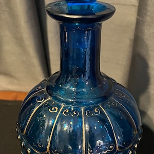 May include: A cobalt blue glass bottle with a faceted blue glass stopper. The bottle has a round body with gold painted details and a gold rim.